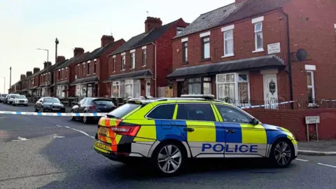 A yellow, blue and white checked police car is parked on a street. Blue and white police tape is cordoning off the street. The street is empty. 