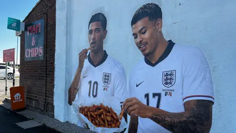 A photographic quality mural of Jude Bellingham on the left and Morgan Rogers on the right eating from a shared bag of chips. It is on a white wall next to a Fish and Chip shop sign
