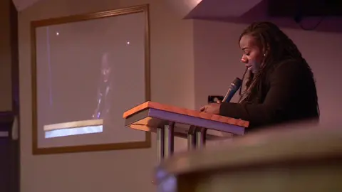 Women standing at a lectern addressing the vigil. She is holding a microphone and a screen broadcasting her address can be seen in the background.