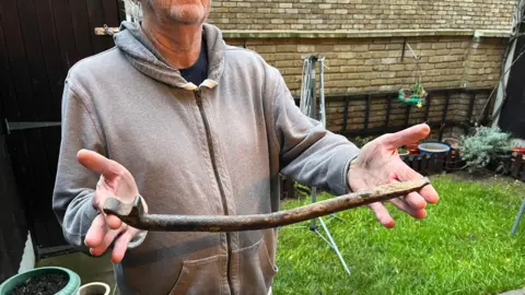 LDRS Torso and arms of a man holding a crowbar. He is wearing a grey hooded top, is standing in a grassed area with flower pots and his head has been cropped out of the image.