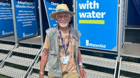 Man with gilet wearing sun hat and lanyard smiles at camera in front of metal steps leading up to a block of festival toilets