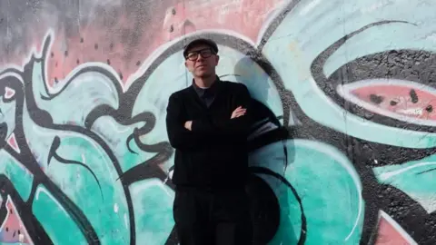 University of Birmingham John Munro stands against a wall on which a colourful image has been spray painted. He is wearing a hat and glasses with a dark shirt, coat and trousers. He has his arms folded. 