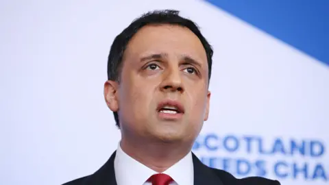 Anas Sarwar photographed at a press conference, wearing a black suit, white shirt and red tie. The photograph is a close-up shot of his head and neck.