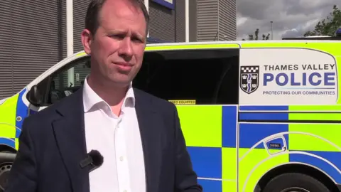 BBC Matthew Barber, with folded arms, is looking at the camera wearing a grey suit and a striped tie.