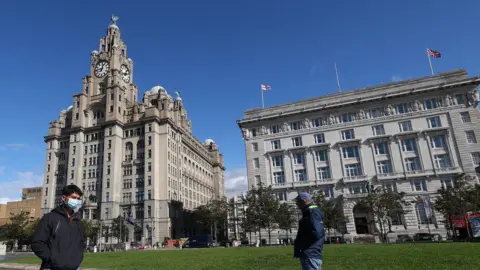 PA Media Two men walk in opposite directions past the Royal Liver Building, a grey stone box shaped structure with two clock towers, each crowned with a statue of the famous Liver Birds. 