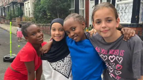 Four young girls pose arm in arm for the camera on the street, where toys can be seen in the background