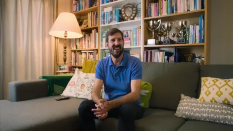 King Putt A man wearing a blue polo shirt and dark trousers is sitting on a grey sofa. There are several pillows next to him and a book shelf and lamp behind him.