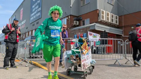 John is kitted out in green with a green wig, a green Macmillan long sleeved running top, green shorts, socks and trainers and an oversided green sponge hand. He pushes a decorated pushchair.