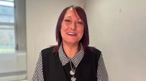 Claire Curran is smiling. Her shoulder length hair is styled in maroon. She's wearing a black vest jumper with a black and white blouse underneath and heart shaped pendants are around her neck. She's standing in a room. 