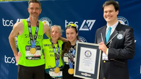 Guinness World Records Two women in running gear and medals, and a man in running gear and a medal, standing and smiling next to a man in a suit holding up a framed certificate for completing a world record. They're standing outside in front of a branded TCS board.