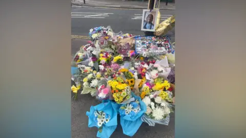 Around 30 bouquets of flowers. all different colours, are arranged on the pavement around a lamp post, on which there is a framed photo of a young man.