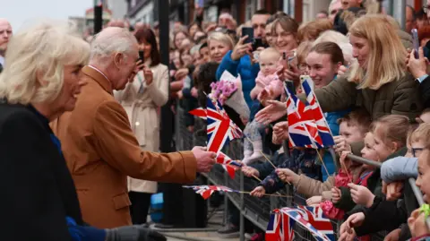 PA Media A number of young people can be seen waving Union flags. 
Some children are smiling as Queen Camilla and King Charles approach them.
King Charles, dressed in a burgundy coat, is pictured about to shake hands with a woman in a green coat. Camilla in a black overcoat has her hand outstretched to greet people in the crowd.