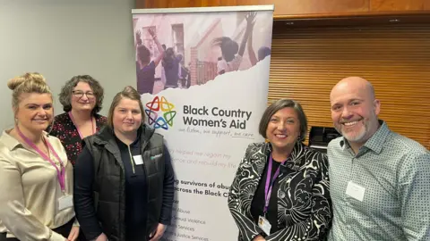 Four women and a man stand together and smile next to a poster saying Black Country Women's Aid.