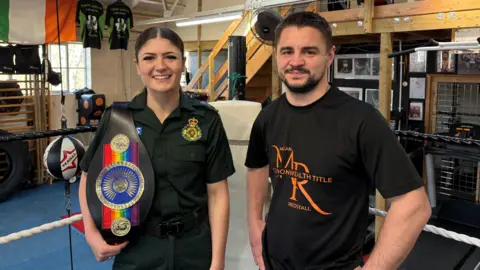 Megan Redstall smiles at the camera while wearing her paramedics uniform and holding her rainbow coloured boxing title belt. Her coach Joe Hughes stands next to her.