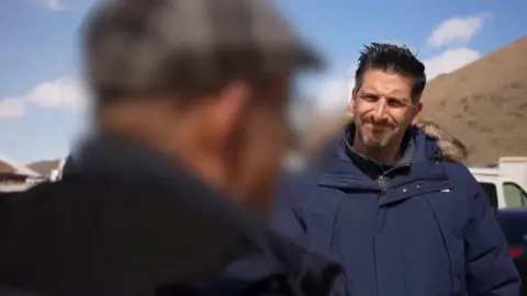BBC correspondent Dan Johnson, wearing navy blue jacket, talking to a man with a blur hiding his face.