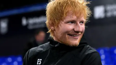 Ed Sheeran sits in Portman Road stadium. He looks away from the camera while smiling. He has long red hair and a red beard. He wears a black rain coat.