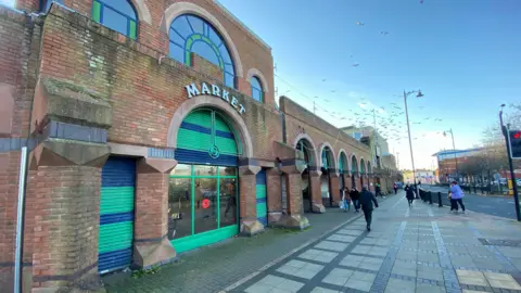 A tall brick building with a sign reading 'market' over the archway window, with green shutters, and a pavement on the right hand side of it with pedestrians walking on a crisp winter day