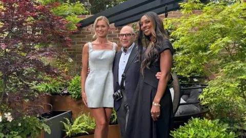 Jen Donnelly Two women in dresses are stood in the garden, an older man wearing a shirt and blazer is stood between the with his arms around theirs waists