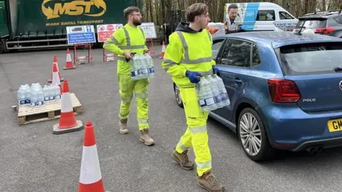 Men in dayglo yellow overalls hold crates of bottled water  