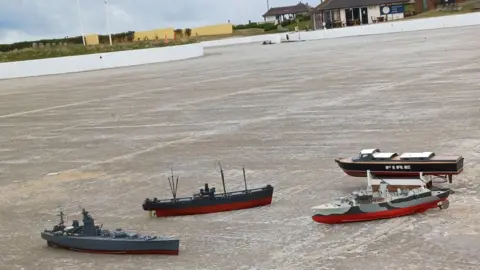 Four model boats laying on the concrete of the empty boating lake in Coatham, Redcar. The structure has a curving shape and is near the sea. The Boathouse and Terrace cafe is visible in the background.
