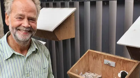 University of Cambridge Dr Mike Rands is smiling at the camera standing in front of two wooden bird nesting boxes. He has greying hair and facial hair and is wearing a striped shirt.
