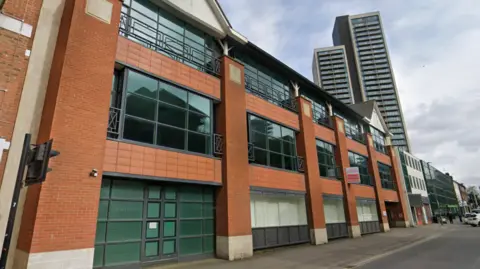 A red-brick terraced building with large windows is spread across three visible floors. A pavement and road with double yellow lines sits at the front of the building. Multi-storey tower blocks and a cloudy, grey sky peak out above the building's roof.