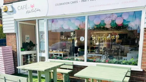 Milliefox Cakes The outside of a cafe with "Cafe at Milliefox Cakes" above the door and green wooden tables in front.