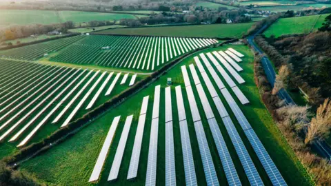 A stock picture of a solar farm. There are rows of solar panels in a field. 