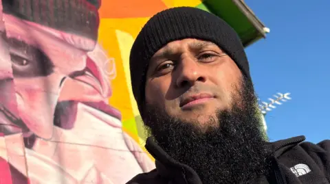 Atiq Rehman standing in front of a brightly coloured mural outside. The shot is taken from below at a slightly skewed angle, looking up and to the left of frame. He is on the right of centre of the frame, wearing a black tight fitted knitted hat, black fleece jacket, which is unzipped, and a black top. He has a long black beard and is looking directly down the camera. The mural behind him shows a man – who is also Atiq – smiling and leaning forward, though he is distorted by the shape of the building. The sky in the top right corner is bright blue and cloudless.