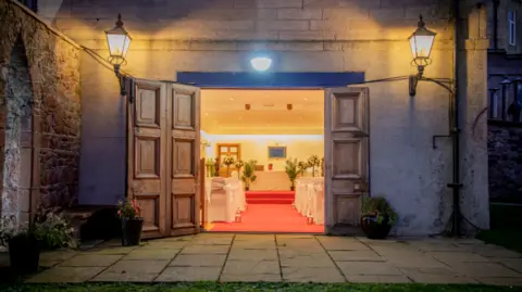 Graham and Sibbald An open wooden door reveals a wedding interior, with white satin furnishings and a red carpet