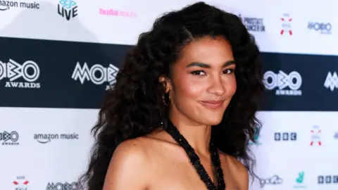 Reuters Olivia Dean wears a black sparkly strappy dress. She is dark brown hair and wears hoop earings. 
