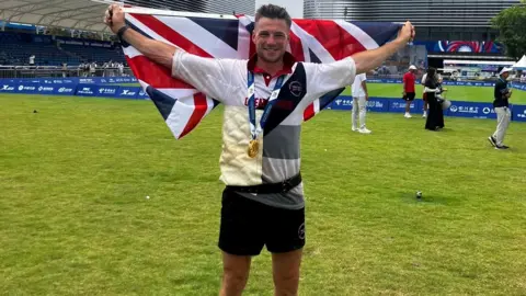 BBC Joe Birch is standing on a sports pitch wearing his gold medal and holding a British flag outstretched behind his arms. He is smiling broadly. 