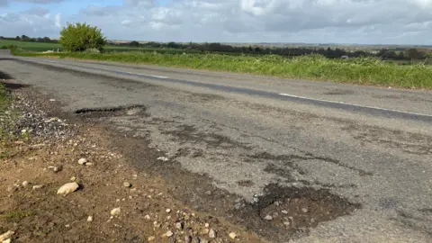 Two potholes on the B4437 outside Burford. It is a small road surrounded by greenery.