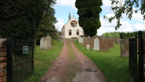 A view up a pathway past gravestones and trees to a white church building with a spire.
