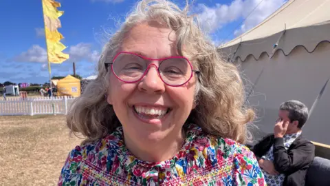 Paige at the Eisteddfod out on the field in sunny weather with a tent to one side and yellow flag behind her. She is laughing and wears a floral blouse and glasses with a pink frame and has curly shoulder-length hair.