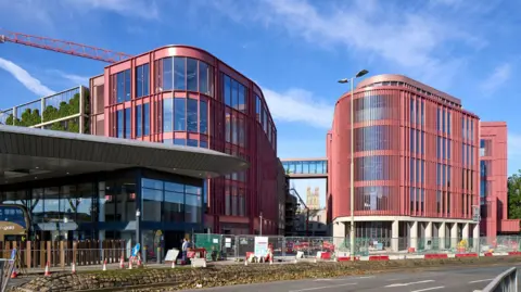 LDRS The Forum in Gloucester as seen from the railway station side of Bruton Way, opposite Gloucester Transport Hub. The Forum is a red brick modern building which has a glass walkway linking to another part of the building. Gloucester Cathedral can be seen in the distance, underneath the walkway.