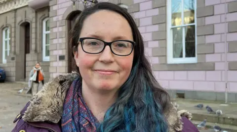 BBC Deputy Louise Doublet in Jersey's Royal Square. She has long dark hair, with blue streaks at the ends. She wears glasses, a purple coat and a patterned scarf. 