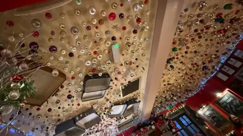 Danny Fullbrook/BBC The ceiling of the pub is decorated with hundreds of baubles. It also is decorated with old open suitcases and a guitar.