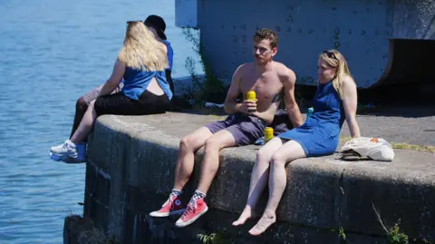 PA Media A man and a women sit on the edge of Bristol Harbour on a sunny day. He is wearing shorts and has his top off, and she is wearing a blue summer dress