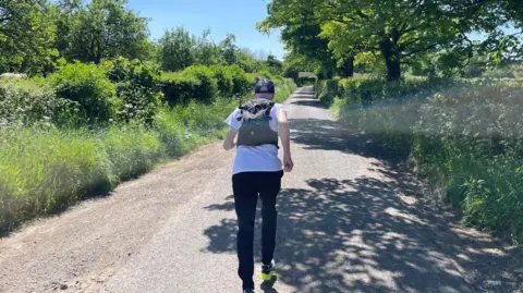 Peter running down a country lane surrounded by trees on a sunny day,. He is wearing a cap, a running vest and dark trousers.