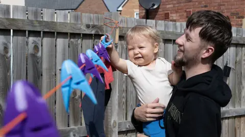 NEYDL Callum Fay is holding his son Archie in his arms as he plays with pegs on a washing line. They're both smiling.