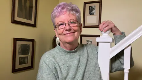 BBC/Victoria Scheer The image shows Maggie standing indoors near a staircase. She is wearing a light green, long-sleeved sweater and has short, light-coloured hair with a hint of purple tone. One hand is resting on the white stair railing, which has a decorative post at the top. The background wall is painted a light yellow shade and features several framed black-and-white photographs arranged in a neat layout. The frames are dark brown and vary in size, with at least four visible in the image.
