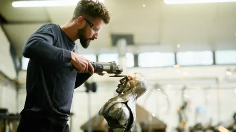 Brian Fell/Adrian Lambert George Fell who has a dark hair and a beard wears a blue sweatshirt and is burnishing a sculpture.