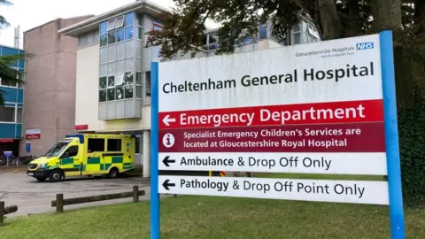 The sign for Cheltenham General Hospital's Emergency Department on a grass lawn in front on the main building, next to a parked ambulance