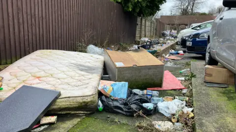 A dirty white mattress is lying on the ground, next to an old wooden wardrobe. They are surrounded by black bin bags and bottles and cans. 