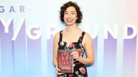 Author Andrea Bartz at an event holding a hardback copy of her novel The Lost Night. She is wearing a black sleeveless dress with a rose pattern  