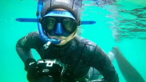 Heather Hamilton/@cornwallunderwater A woman in a wetsuit and a snorkel. She is underwater, holding a camera.