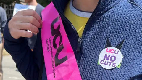 A close-up of a woman wearing a pink banner with the words UCU University and College Union Scotland. The woman is also wearing a sticker which says UCU No Cuts
