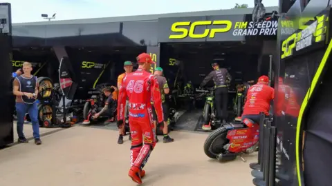 A race track driver in a red race suit with the number '46' on, walks towards a pit full of other men working on various motorcycles. It is a sunny day. 
