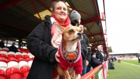 PA Media A Stanley fan holds her dog, with a red jumper, on the side of the pitch before an FA Cup game